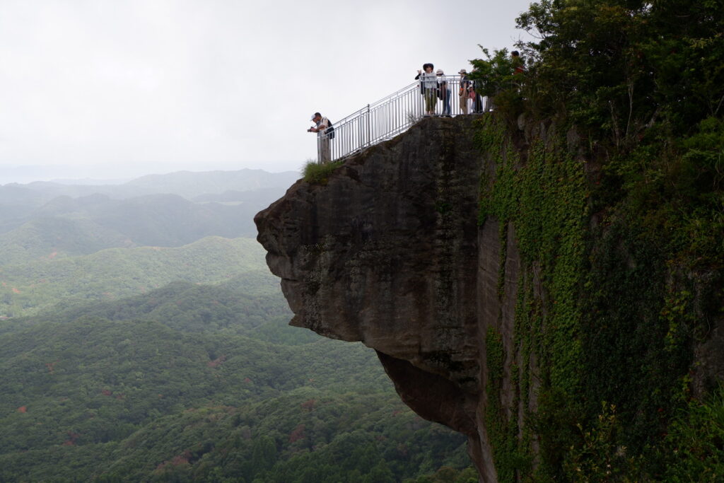 千葉県・鋸山の断崖から突き出した展望台。柵沿いに観光客が並び、眼下に緑の山並みが広がっている