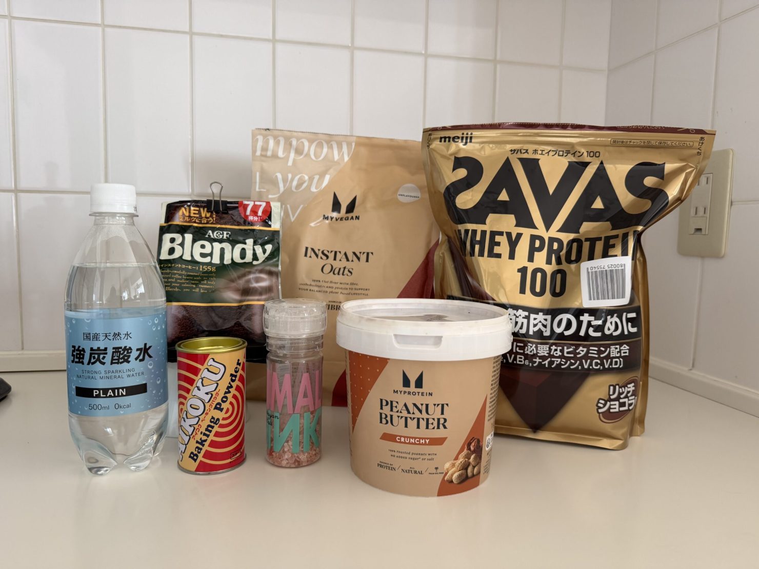 実際に使っている材料を並べた写真。強炭酸水、AGFブレンディのインスタントコーヒー、MYVEGANのインスタントオーツ、明治ザバス ホエイプロテイン100 リッチショコラ、AIKOKUのベーキングパウダー、ピンク岩塩のミル、マイプロテインのピーナッツバター クランチ